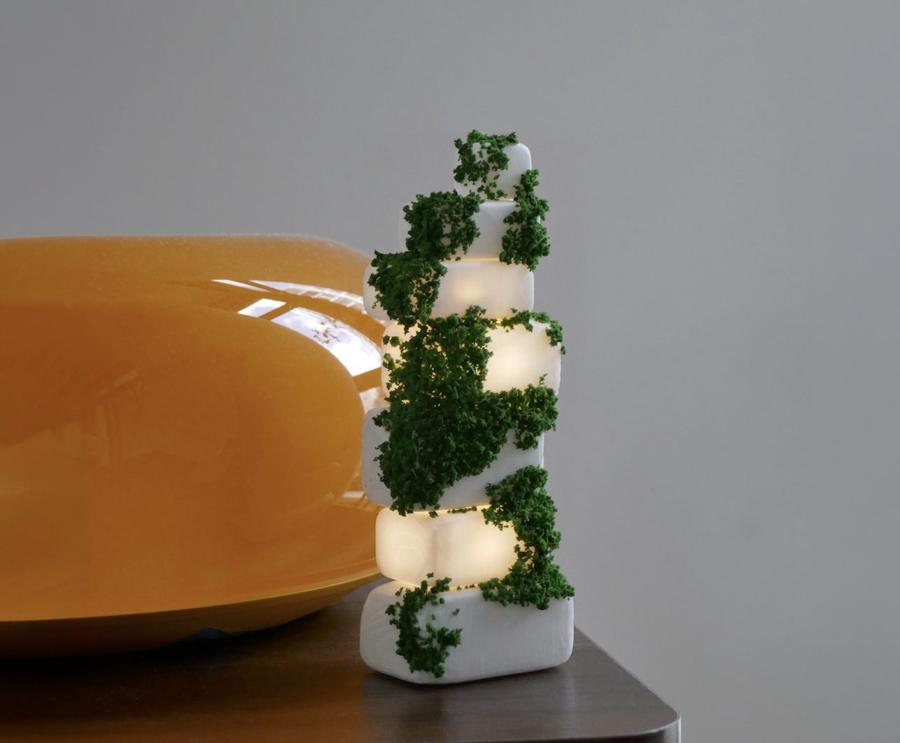Cairn — a 3D-printed white stone-stack object threaded with green moss, glowing from between its blocks